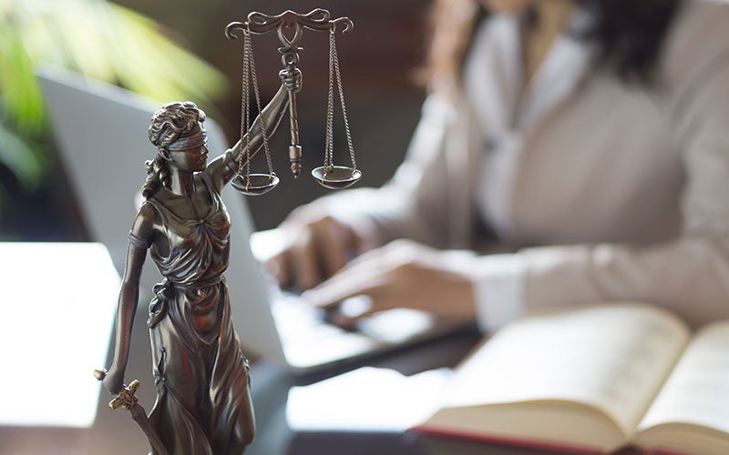A Lady Justice statue with scales and a person typing on a laptop in the background.