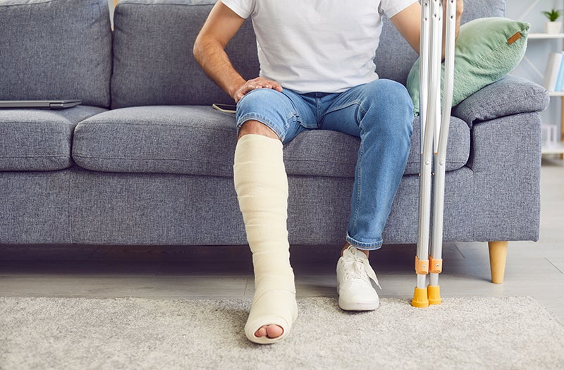 A person with a bandaged leg sits on a sofa next to crutches.