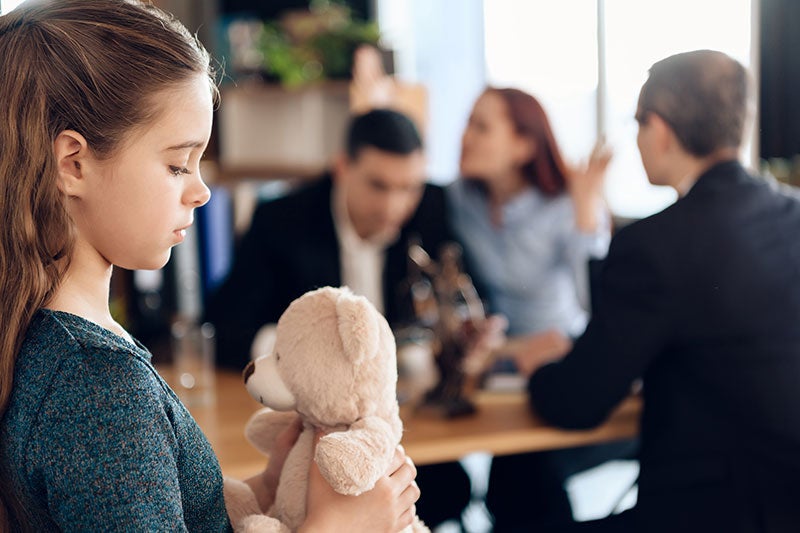A sad young girl holds a teddy bear as her parents argue in the background during what appears to be a divorce proceeding.