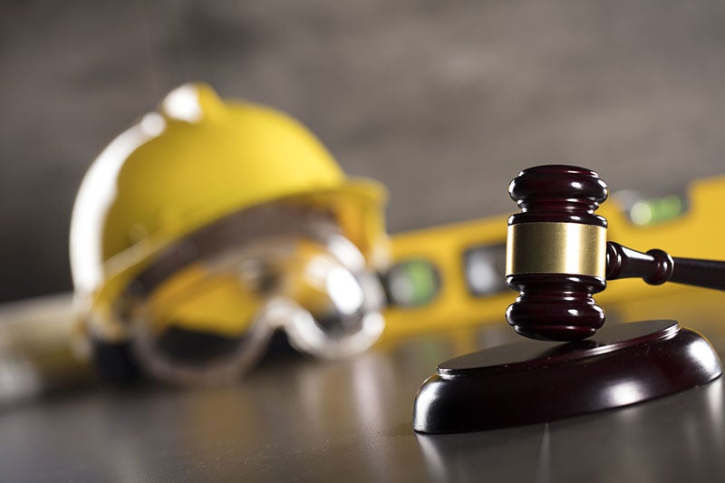 A wooden gavel in the foreground, with a yellow hard hat and safety goggles