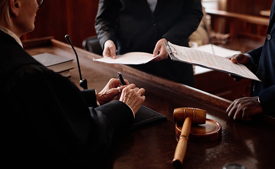 A person in a black robe sits at a wooden bench holding a pen. Legal documents and a gavel are nearby.