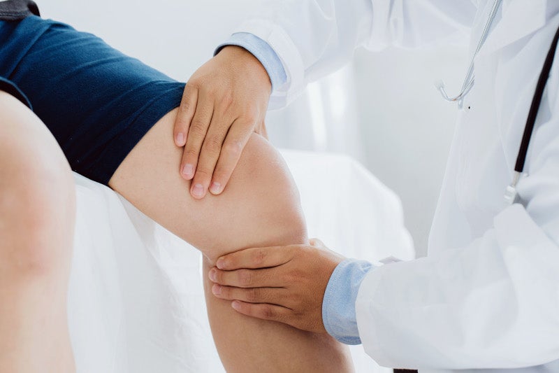 A doctor in a white coat examines a patient's knee by holding their leg with both hands.