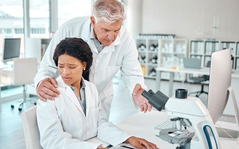 Two scientists in lab coats in a lab. An older man stands, hand on the shoulder of a younger, distressed-looking woman, as she works at a microscope.