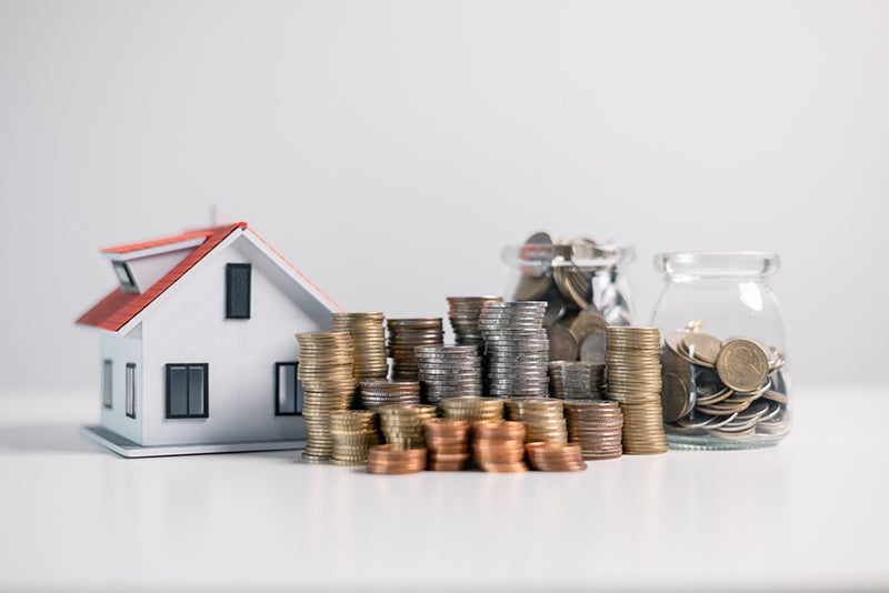A white model house with a red roof stands next to stacks of coins and two jars of coins.