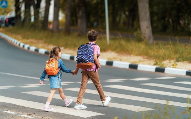 Two children with backpacks, holding hands, walk across a crosswalk.